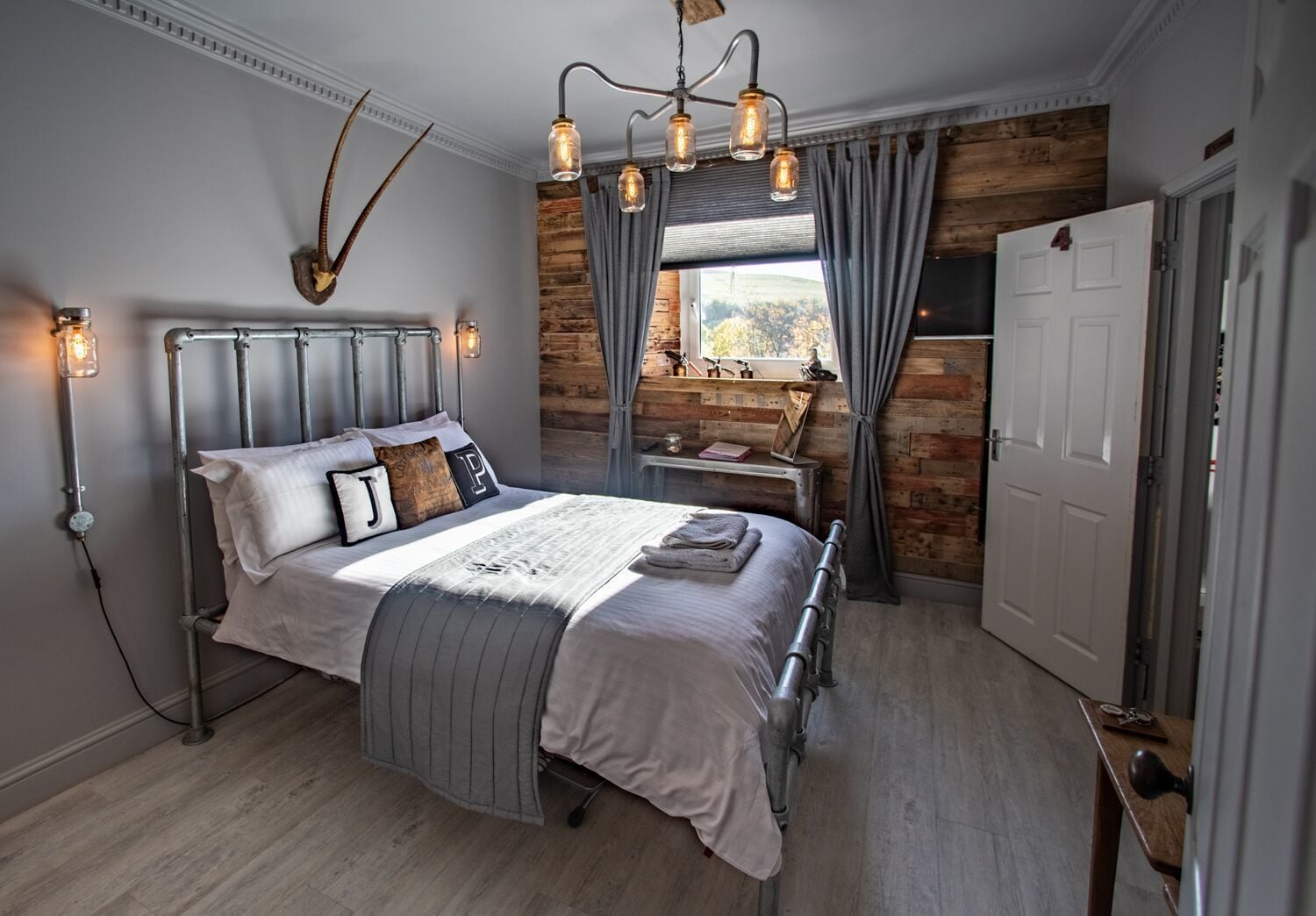 A bedroom with a metal-framed bed, a wooden wall, a window with curtains, and a chandelier.