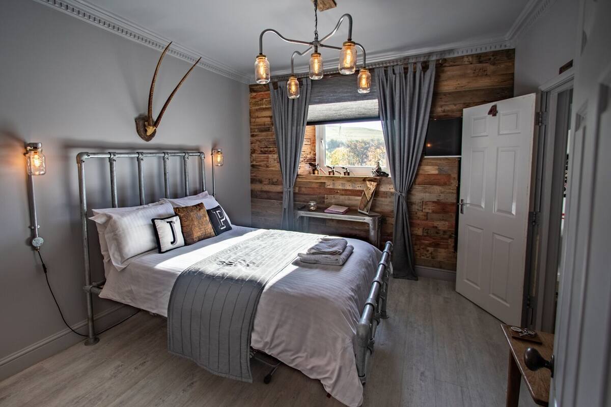 A bedroom with a metal bed frame, a large window with curtains, a wooden wall, and a chandelier.