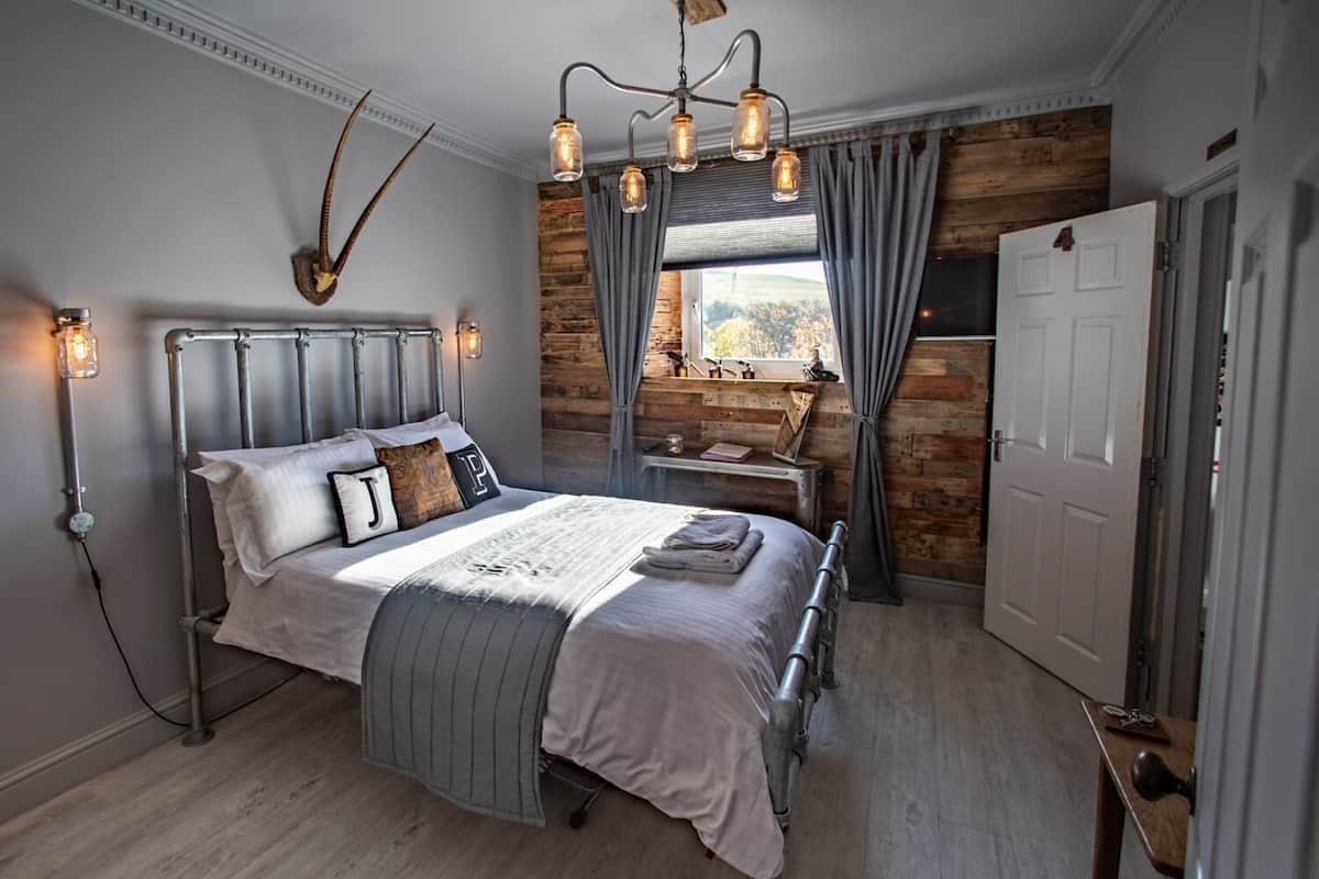 Un dormitorio con una cama de estructura metálica, pared de madera, ventana con cortinas y una araña.