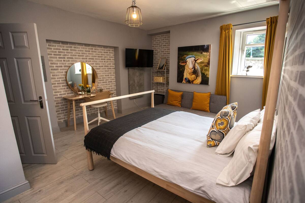 A bedroom with a bed, a desk, a mirror, a TV, and a window with curtains.