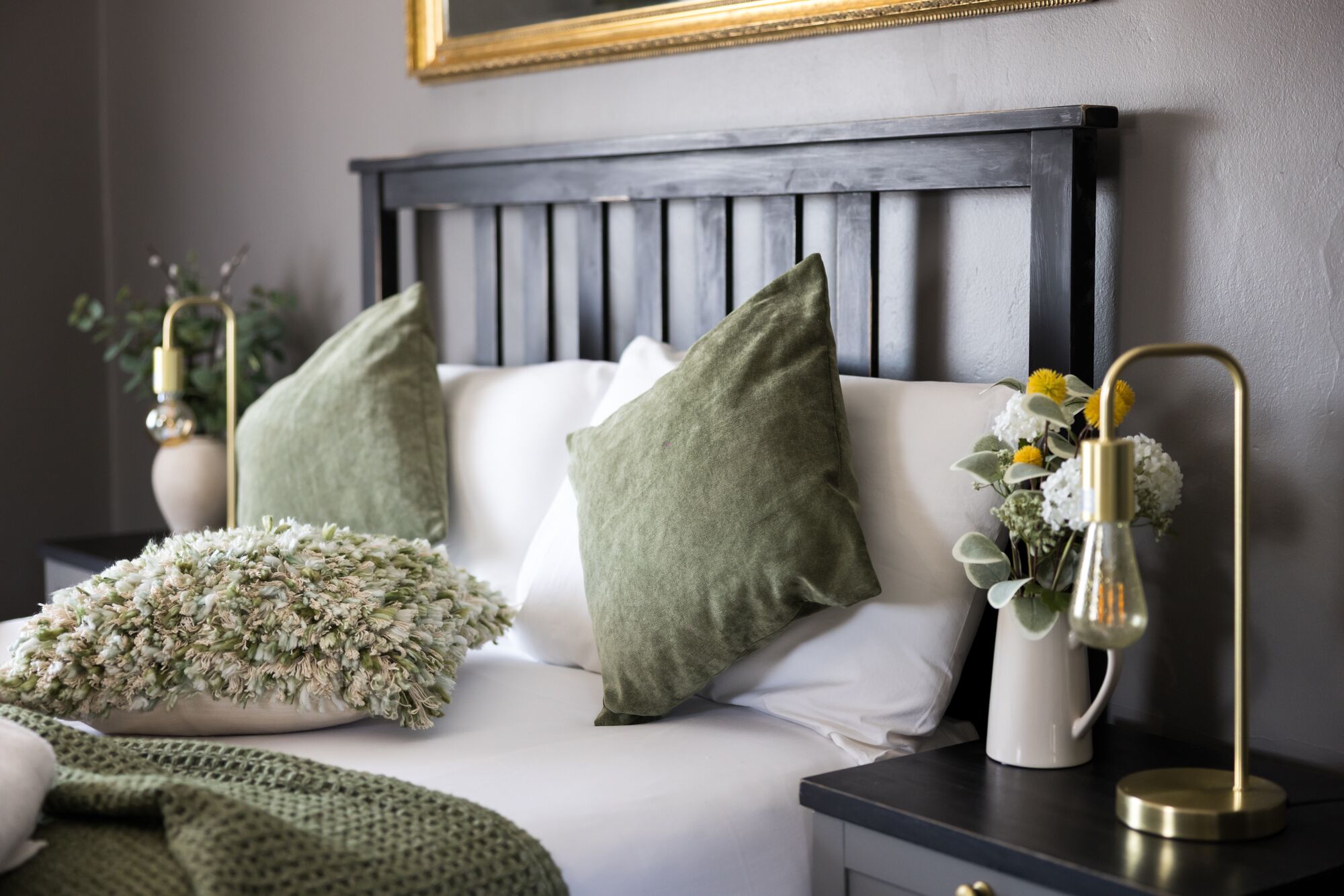 Een net opgemaakt bed met groene en witte kussens, een decoratieve plant en een nachtkastje met een lamp.