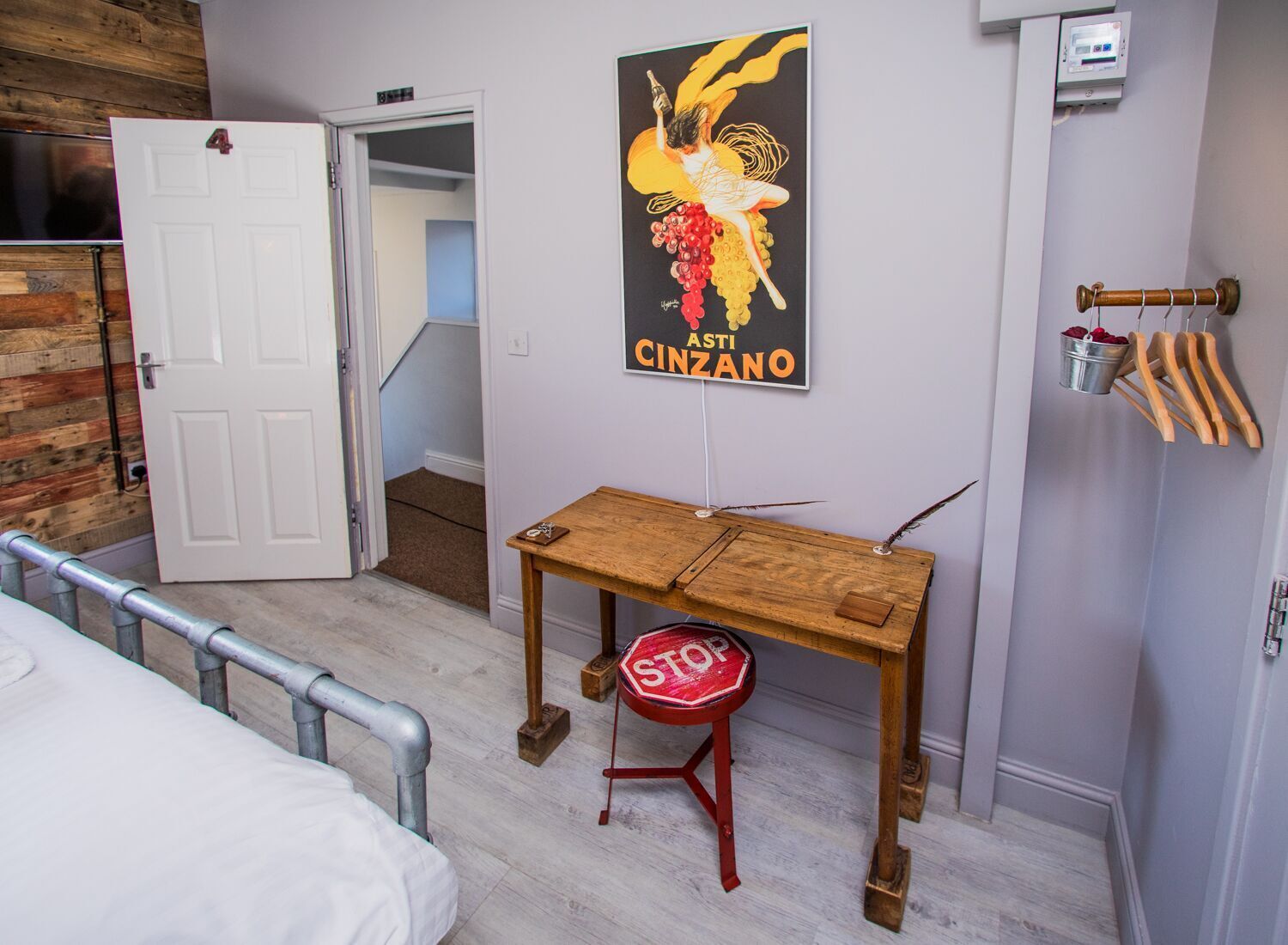 A hotel room with a bed, a wooden table with a red stool, a poster on the wall, and a door leading to another room.