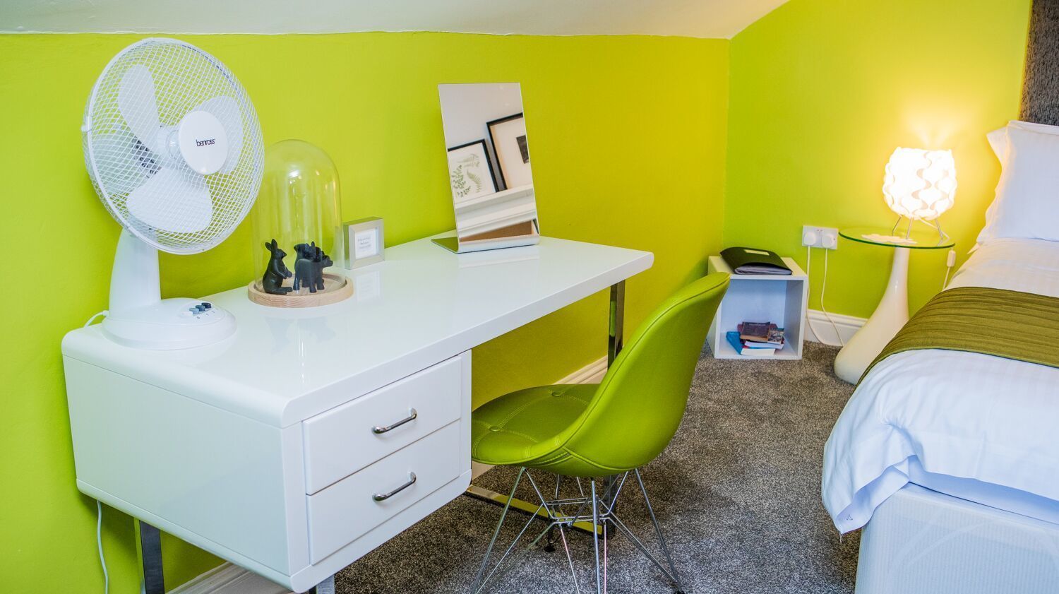 Habitación de hotel con un escritorio blanco, una silla verde, cama, mesita de noche con lámpara y una pequeña estantería con libros y un objeto decorativo.