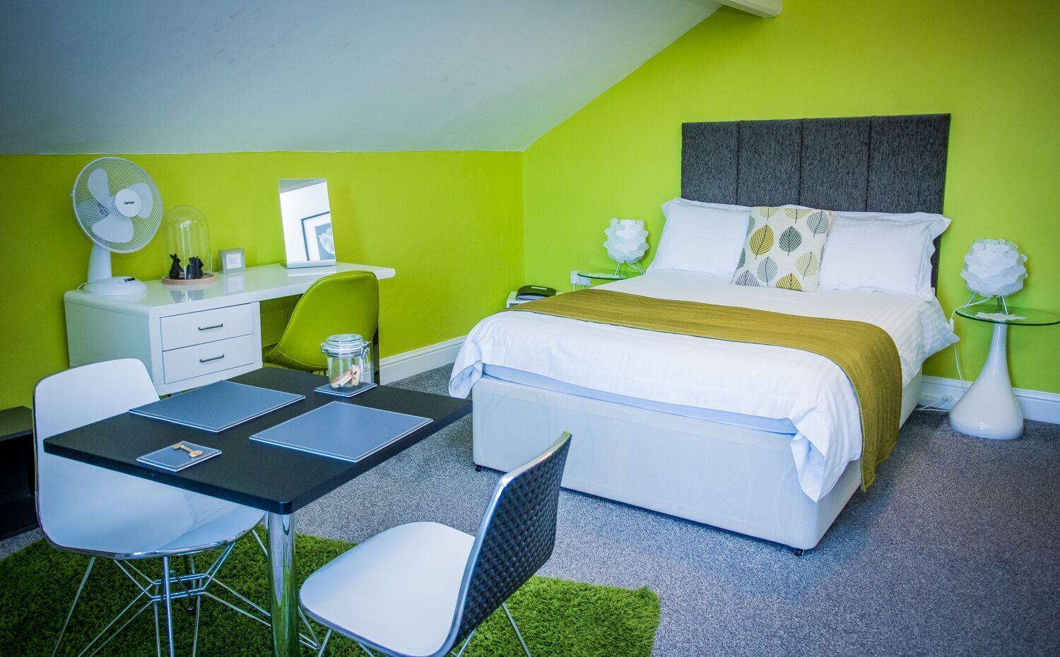 A bedroom with a bed, bedside table, lamp, and a desk with chair and lamp.