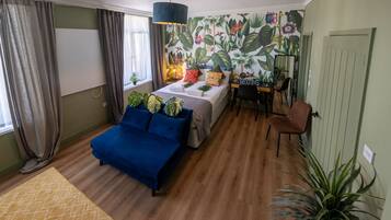 A room with a bed, a blue sofa, a desk, and a chair. There is a window with curtains, a wall with a floral mural, and a plant on the floor.