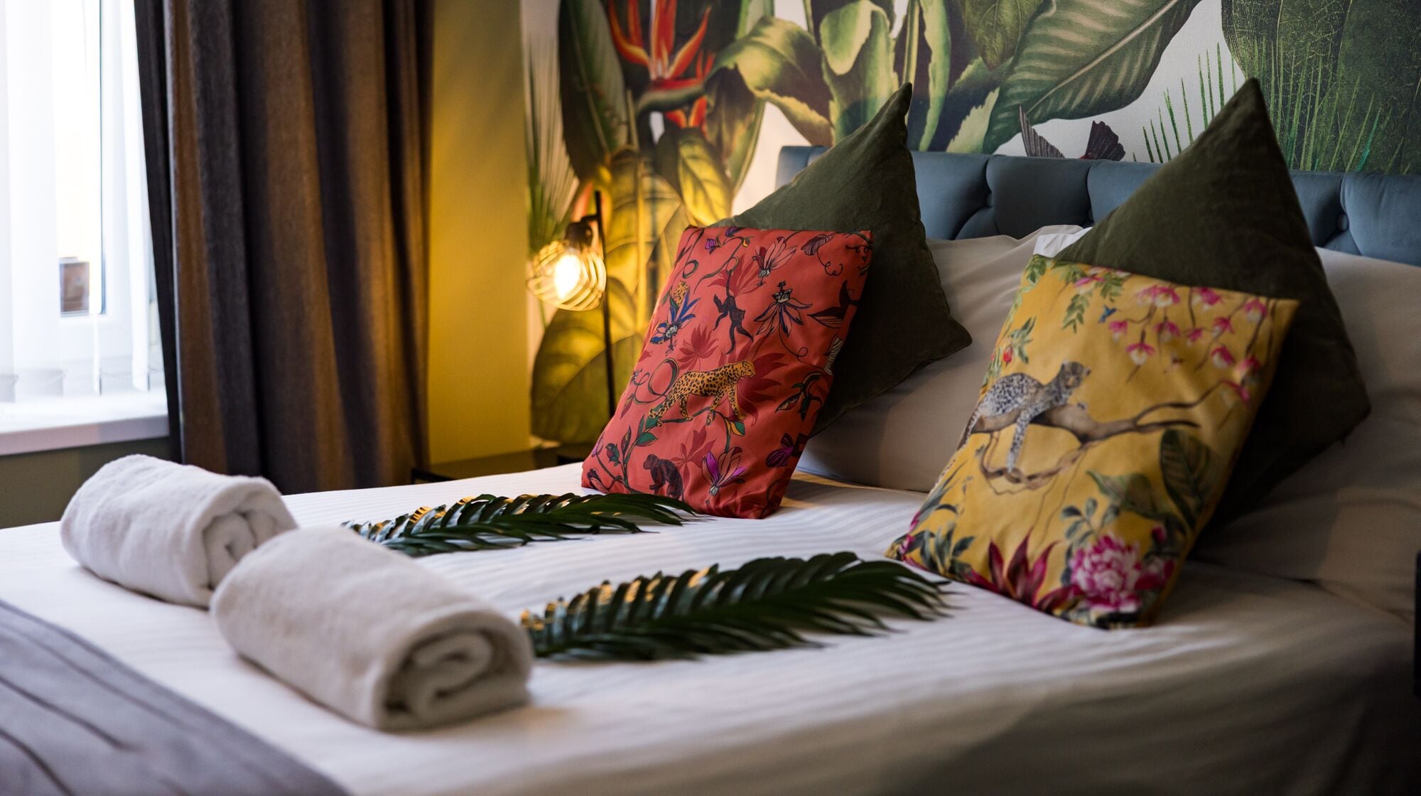 A neatly made bed with rolled towels, two decorative pillows, and a tropical-themed wall mural.