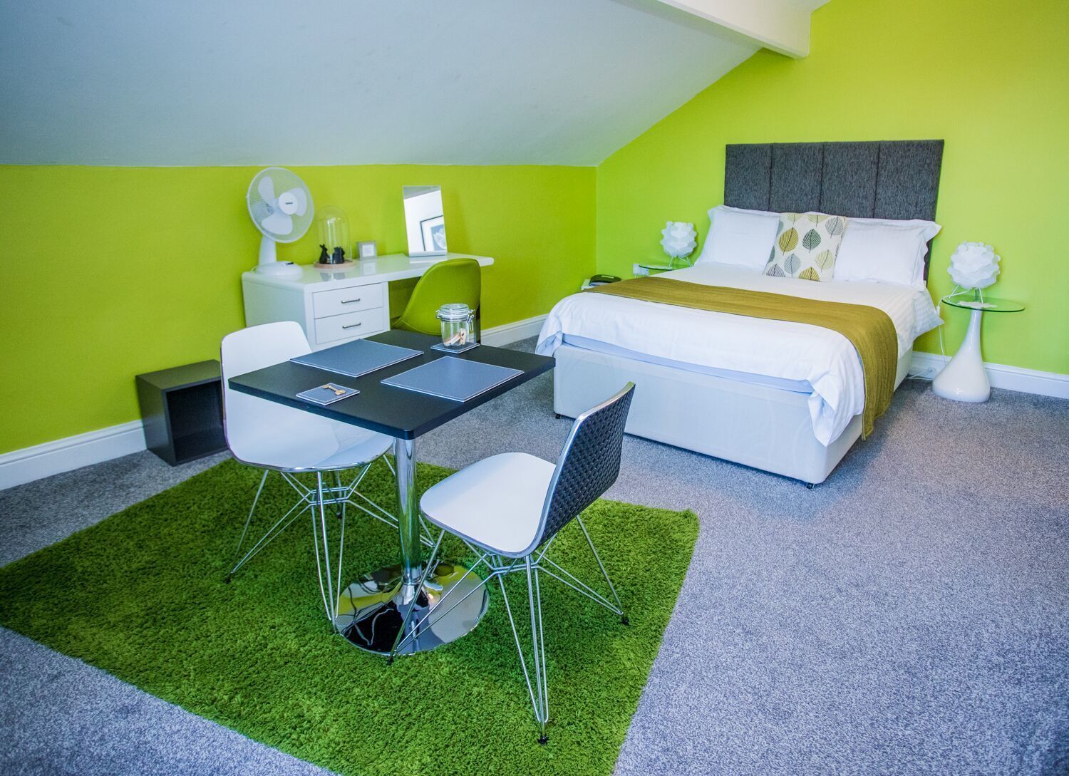 Une chambre à coucher de taille modeste, comprenant un lit, des tables de chevet, un bureau avec des chaises et une moquette verte.
