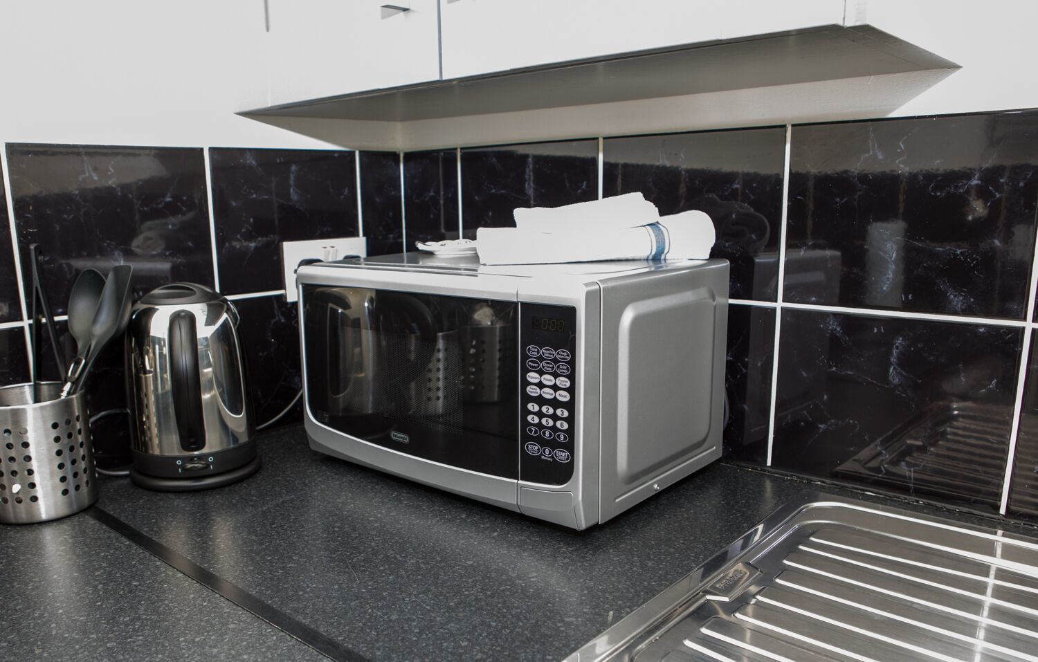 A kitchen with a microwave, kettle, and a stainless steel sink.
