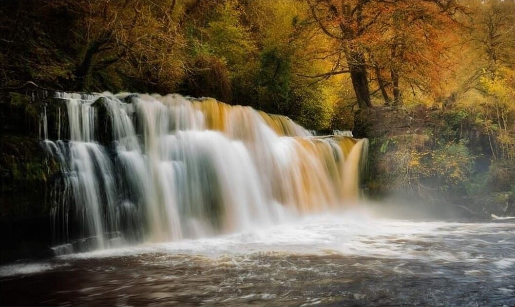 Een waterval in een bebost gebied met weelderige begroeiing en bomen in herfstkleuren.