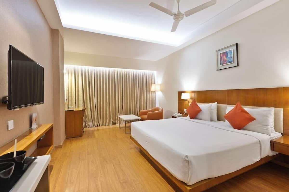 A hotel room with a bed, a TV, a ceiling fan, a desk, a chair, and a window with curtains.
