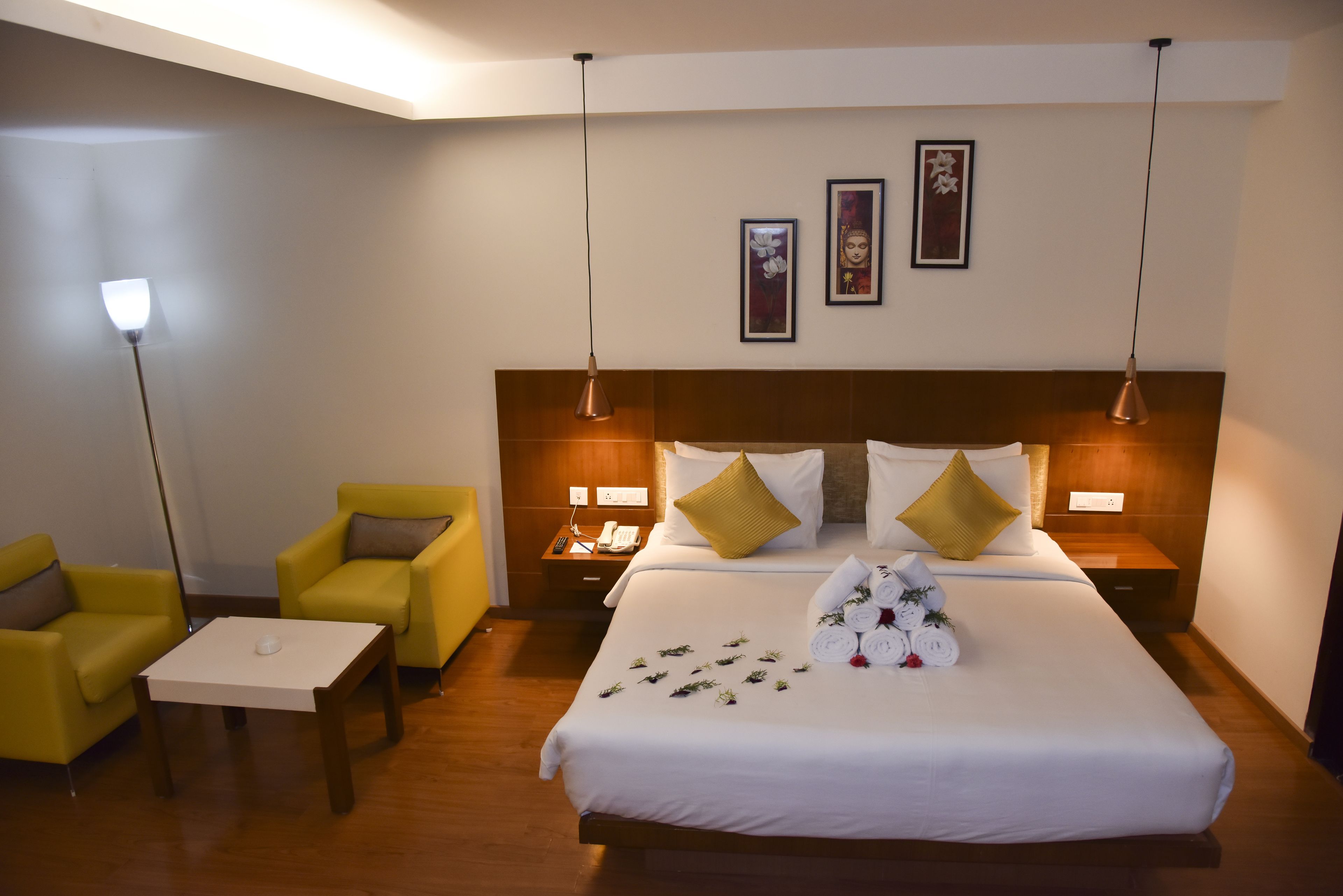 Una habitación de hotel con una cama grande, dos mesitas de noche, una mesita pequeña, dos sillones amarillos y una lámpara de pie.