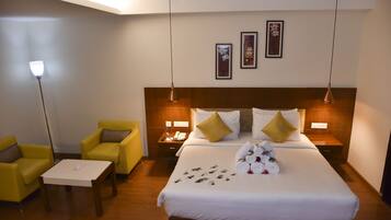 Una habitación de hotel con una cama grande, dos mesitas de noche, una mesita pequeña, dos sillones amarillos y una lámpara de pie.
