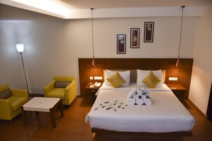Una habitación de hotel con una cama grande, dos mesitas de noche, una mesita pequeña, dos sillones amarillos y una lámpara de pie.