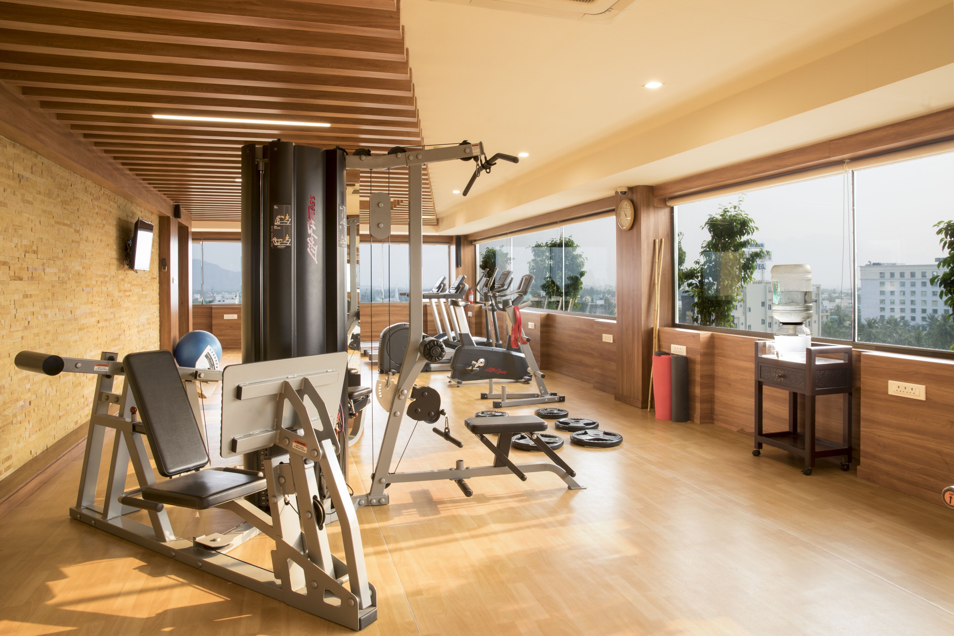 Un gimnasio con diversos equipos de ejercicio, ventanas amplias con vista a la ciudad y suelo de madera.