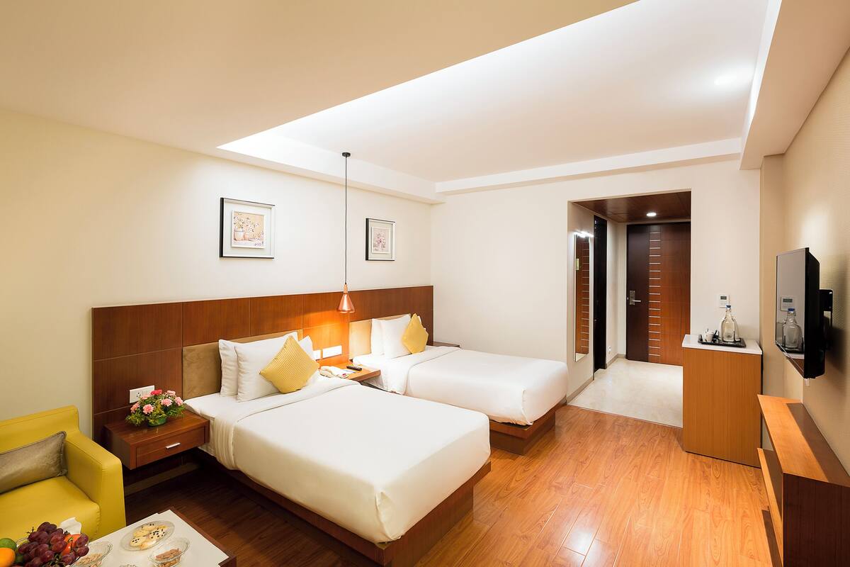 A hotel room with two beds, a desk, a chair, a TV, and a small table with fruit.