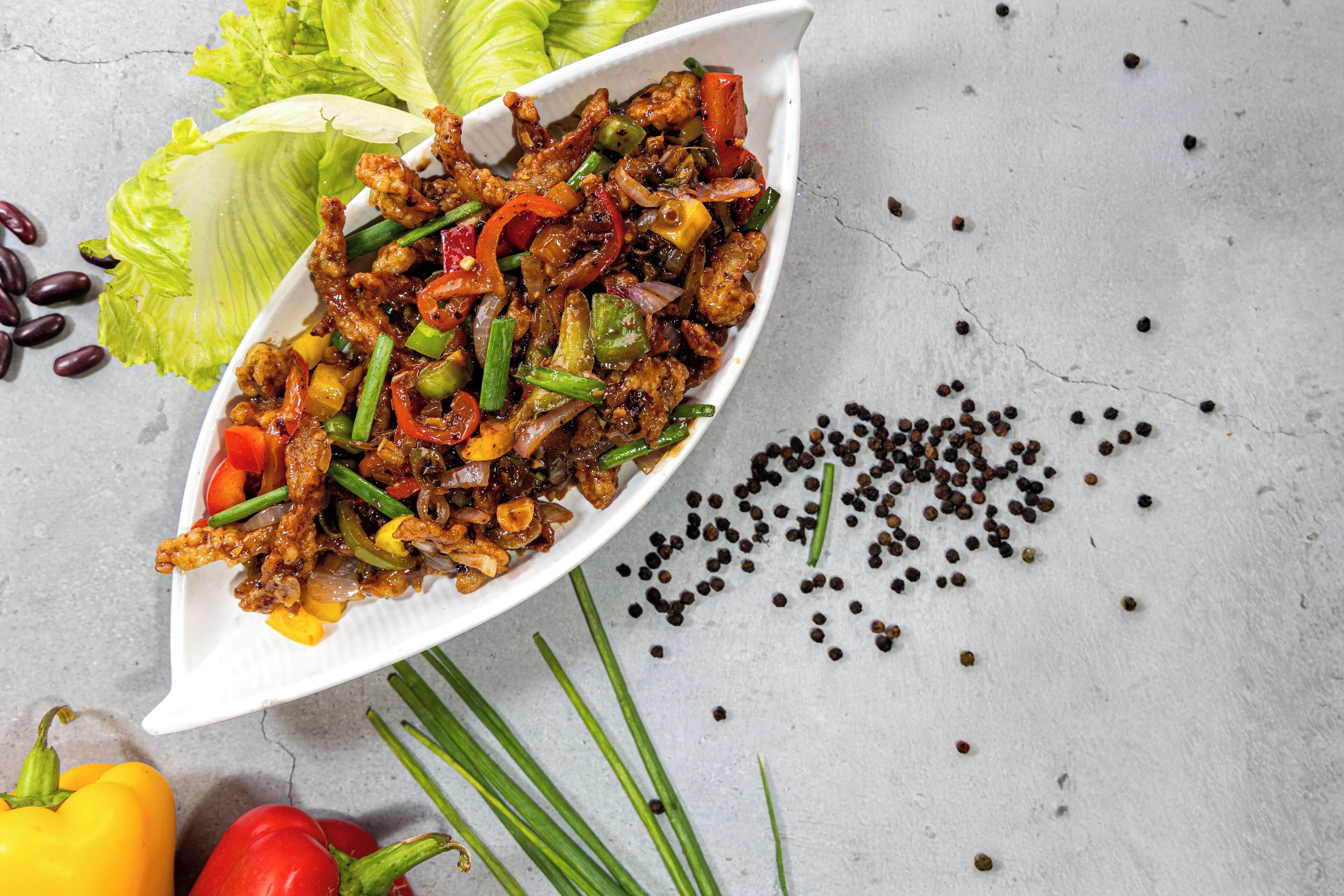 A white dish with a stir-fry containing vegetables and meat, surrounded by scattered peppercorns and whole peppers.