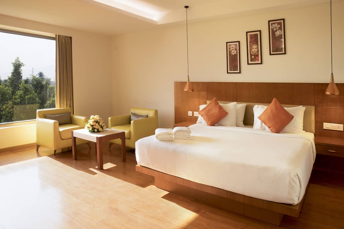 A hotel room with a large bed, two armchairs, a small table with a vase of flowers, and a view of greenery outside.