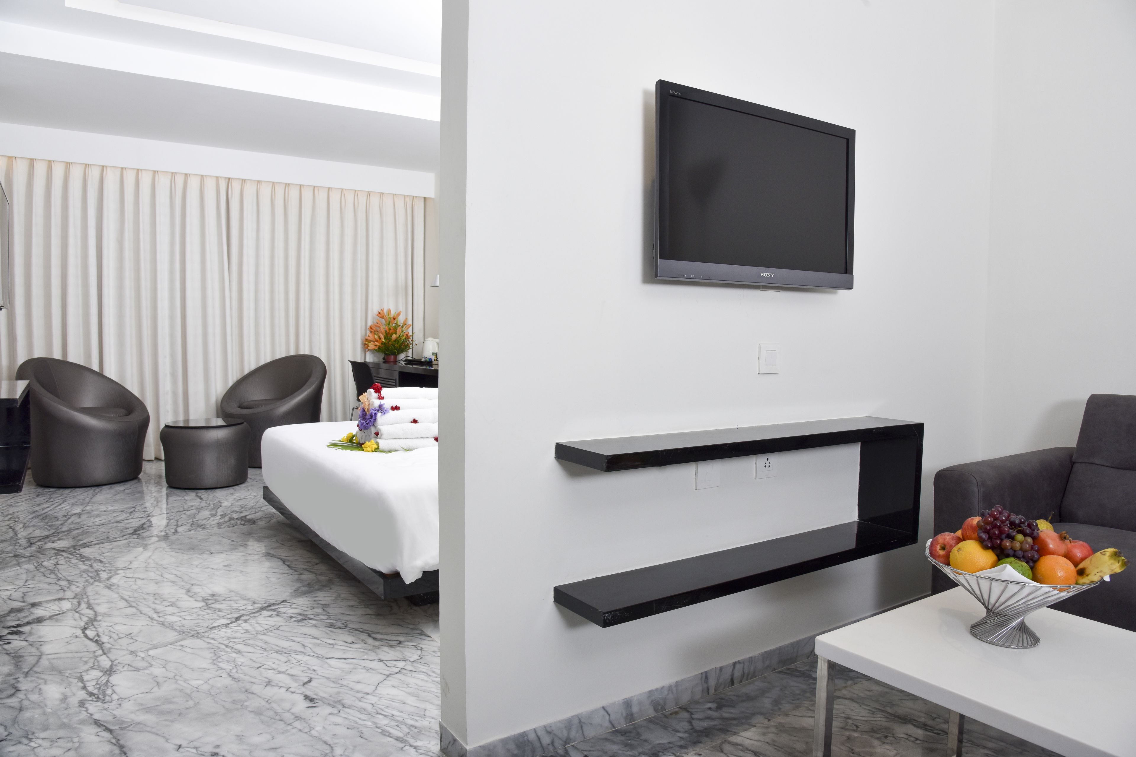 A modern hotel room with a flat-screen TV, a bed, two chairs, a small table with a fruit bowl, and a marble floor.