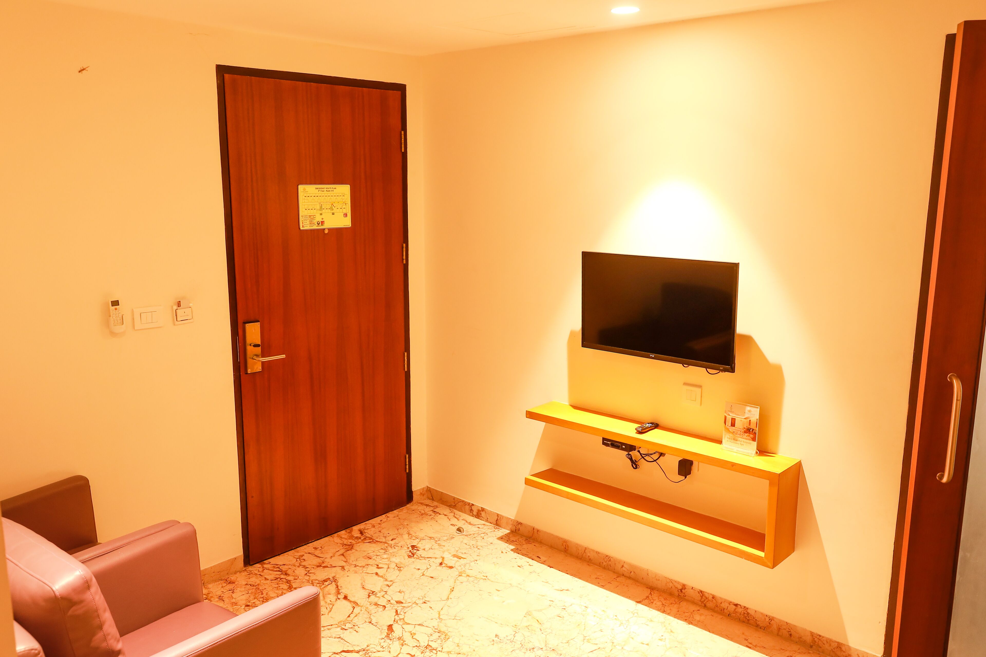 A hotel room with a wooden door, a flat-screen TV mounted on a wooden shelf, a marble-patterned floor, and a small seating area with a chair.