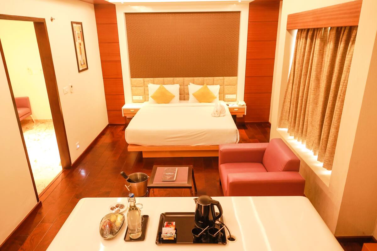 A hotel room with a large bed, a sitting area with a red chair, a bedside table with a tray of breakfast items, and a large window with curtains.