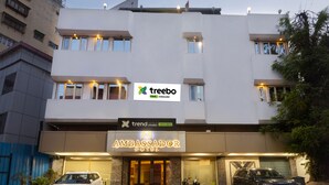 Un edificio moderno con un cartello per 'Ambassador Hotel' e il marchio 'Treebo'. Due auto parcheggiate davanti.