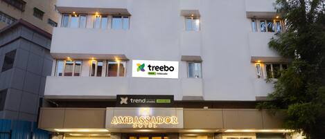 A modern building with a sign for 'Ambassador Hotel' and 'Treebo' branding. There are two cars parked in front.