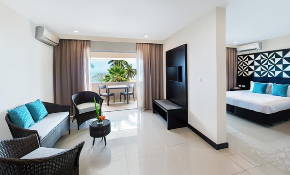 A modern hotel room with a balcony, a flat-screen TV, a bed with blue bedding, and wicker furniture. - Tanoa International Dateline Hotel (Nuku'alofa)