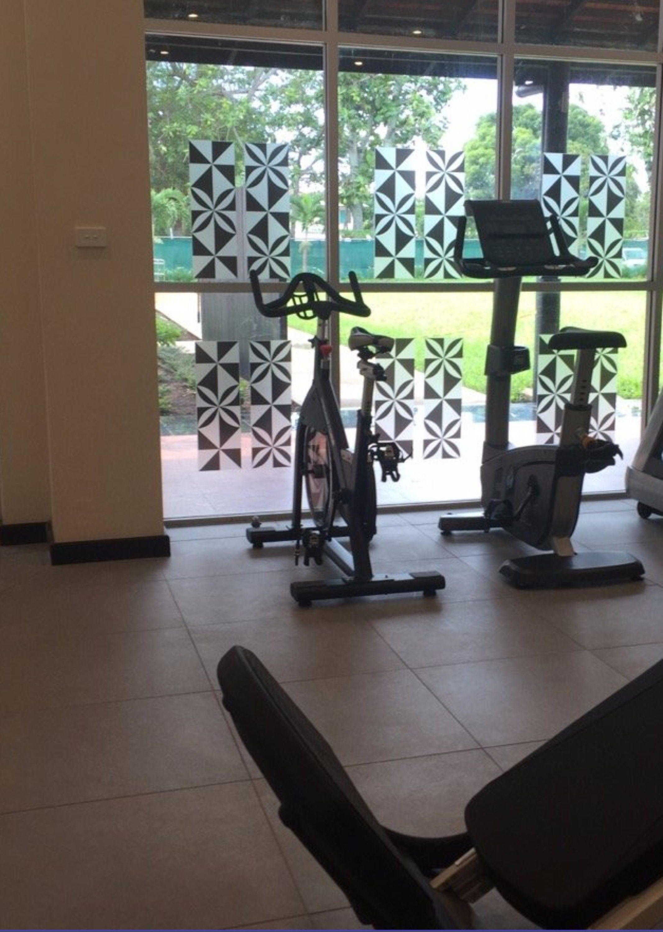 Un gimnasio con bicicletas estáticas y una máquina de remo, amplias ventanas con persianas decoradas y un banco.