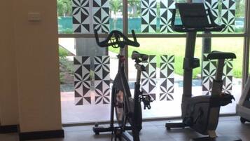 Un gimnasio con bicicletas estáticas y una máquina de remo, amplias ventanas con persianas decoradas y un banco.