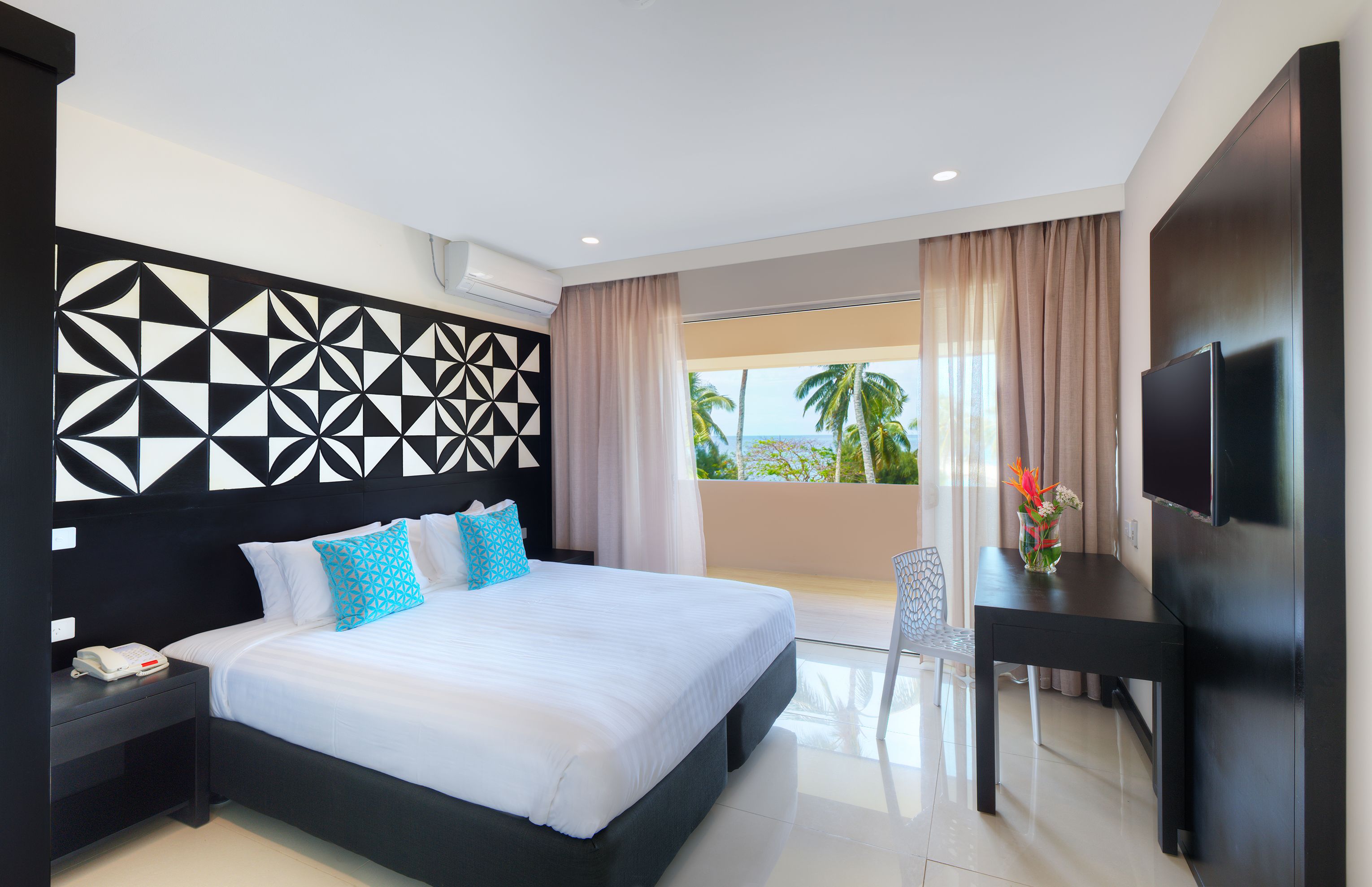 A modern hotel room with a large bed, a desk, a flat-screen TV, and a balcony with a view of palm trees.