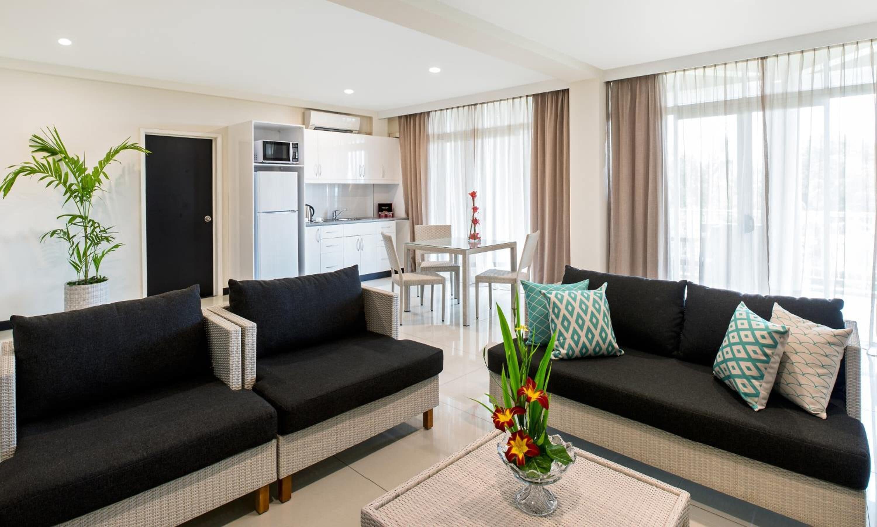A modern living room with a black sofa set, a dining table with chairs, a kitchen area, and a large window with curtains.