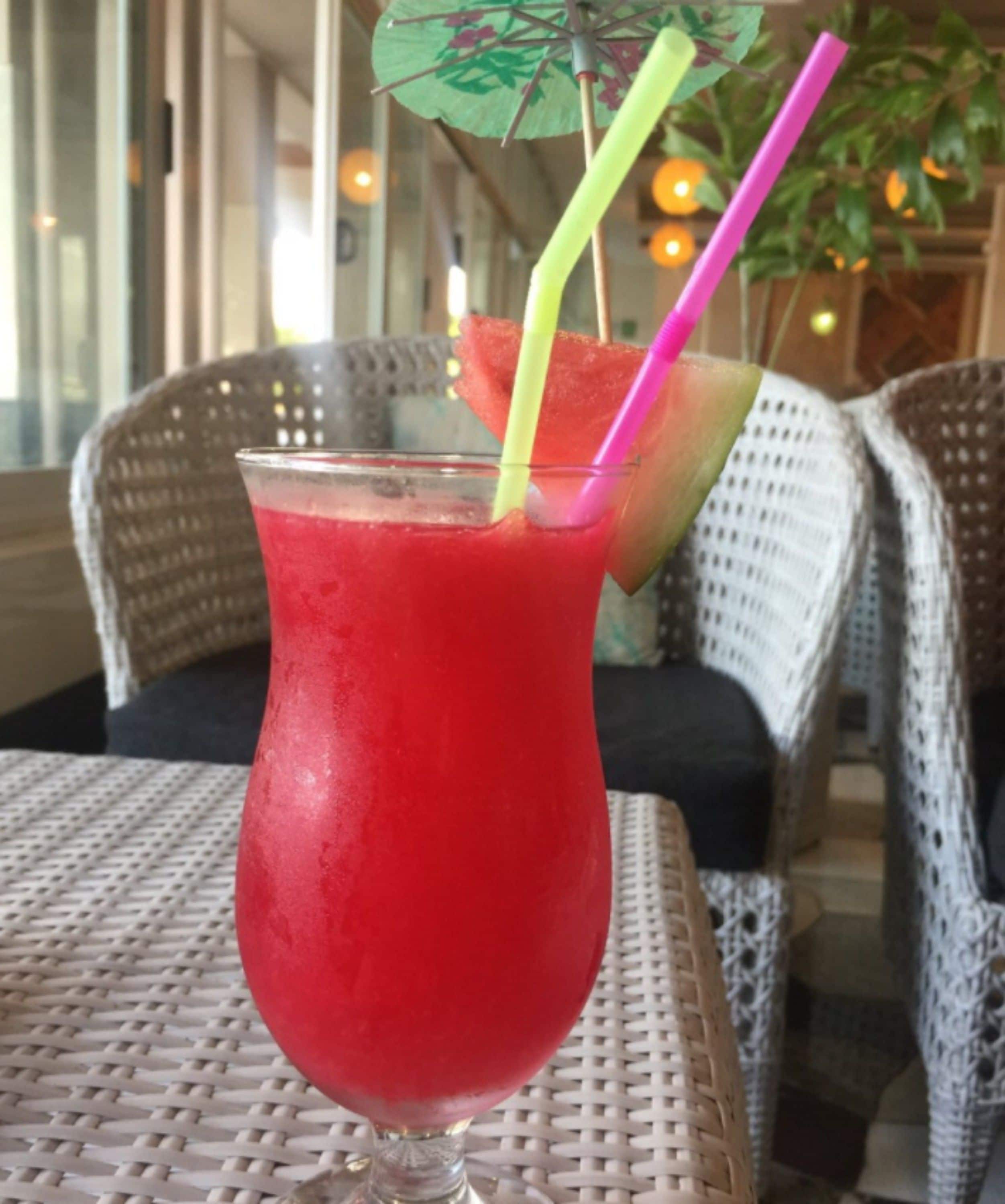 Un bicchiere di bevanda rossa con un ombrello verde e due cannucce su un tavolino in vimini.