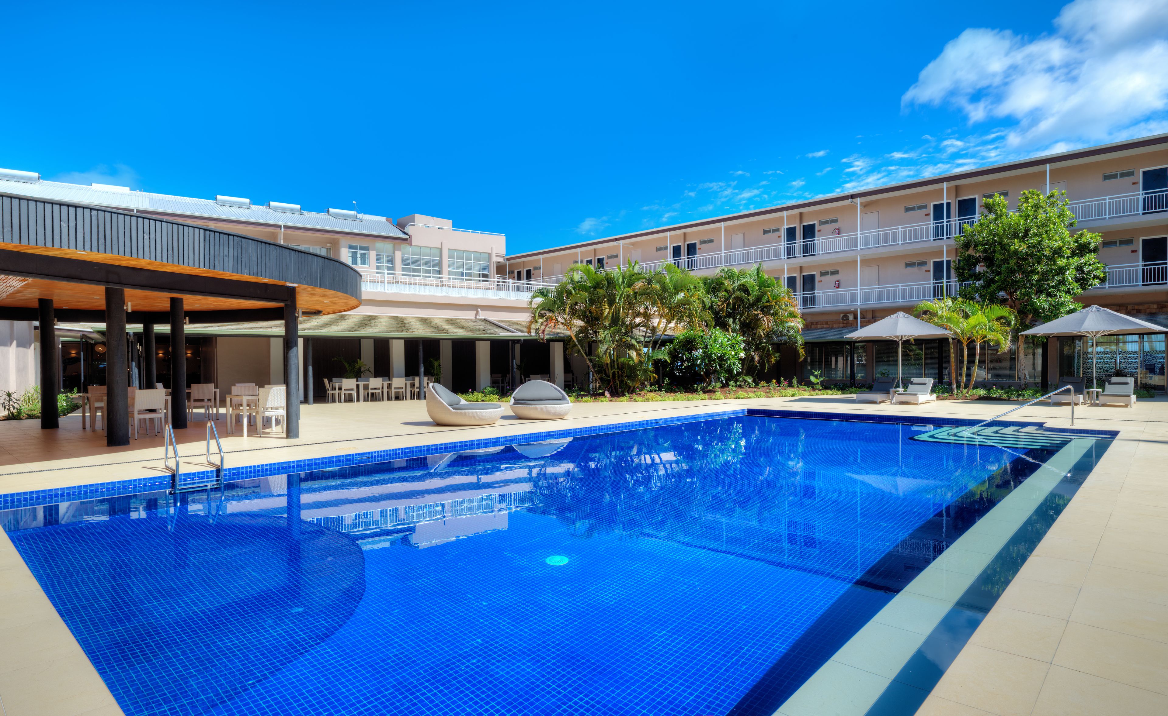 Una piscina con sillas de descanso y sombrillas, rodeada por un edificio con varios balcones y un área de patio techada.