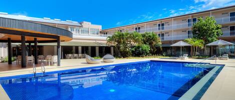 Una piscina con sillas de descanso y sombrillas, rodeada por un edificio con varios balcones y un área de patio techada.