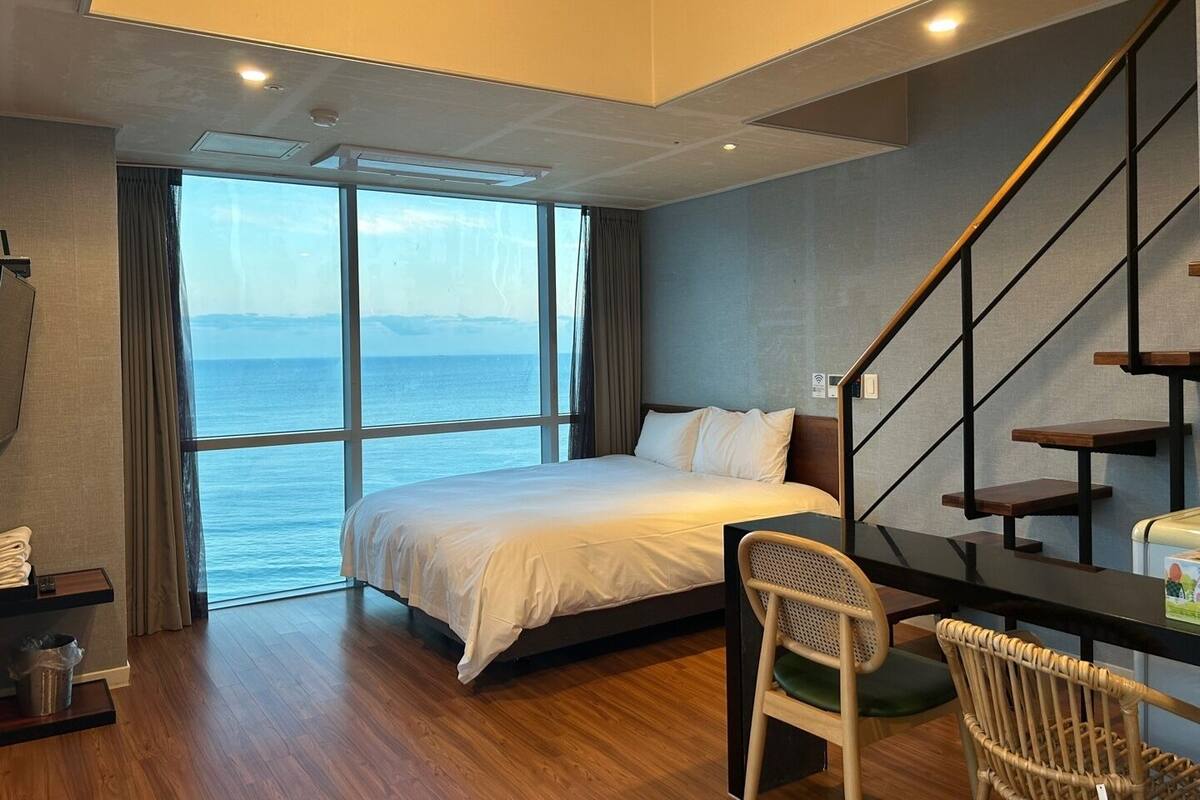 A hotel room with a bed, a chair, a desk, and a view of the sea.