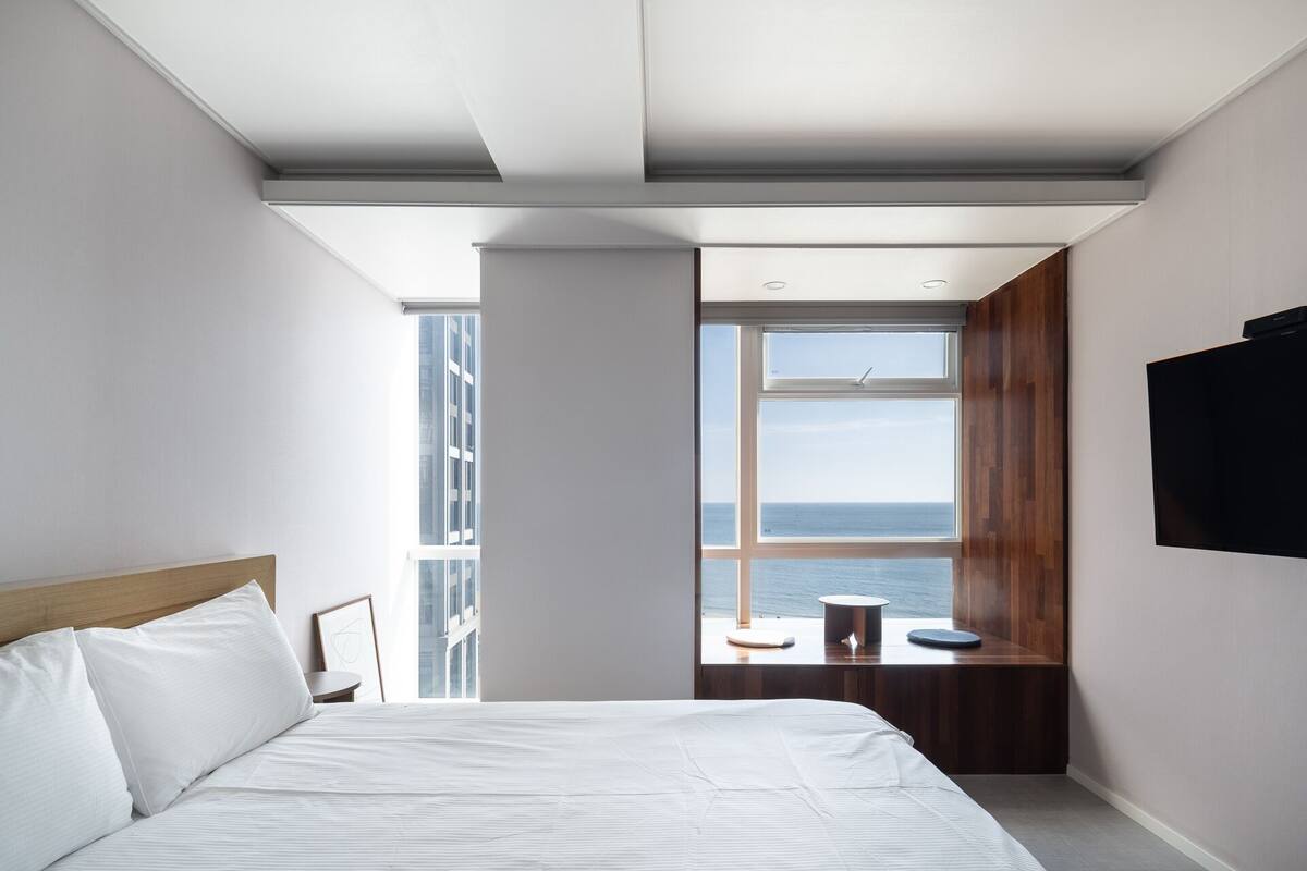 A modern bedroom with a large bed, a view of the sea through a window, and a flat-screen TV mounted on the wall.