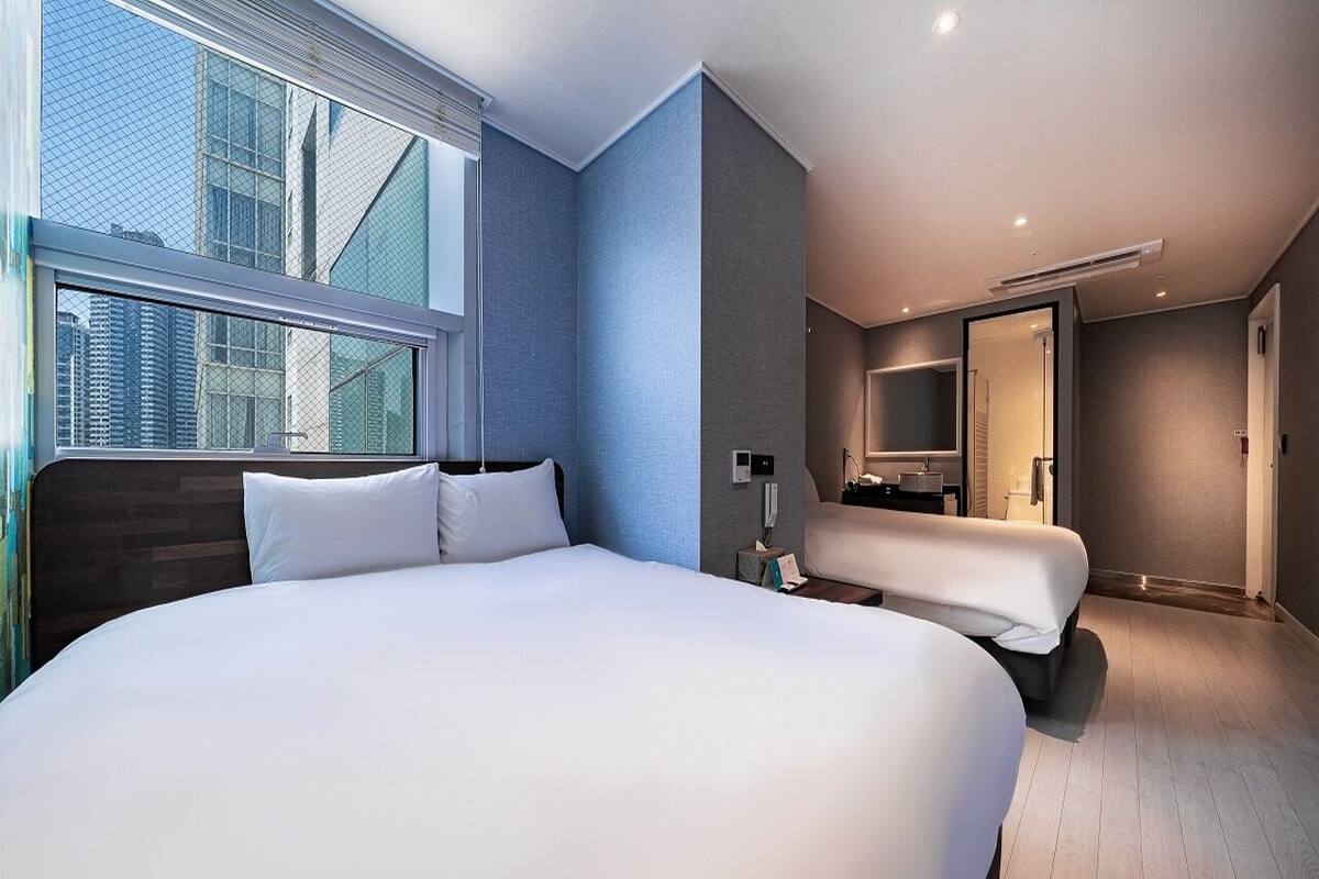A modern hotel room with a large bed, a bedside table, a mirror, and a view of the cityscape through the window.