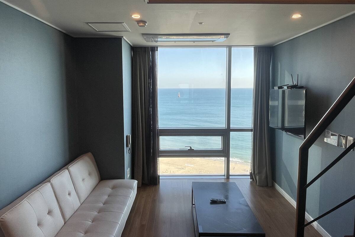 A modern room with a sofa, a small table, a TV mounted on the wall, and a large window offering a view of the sea.