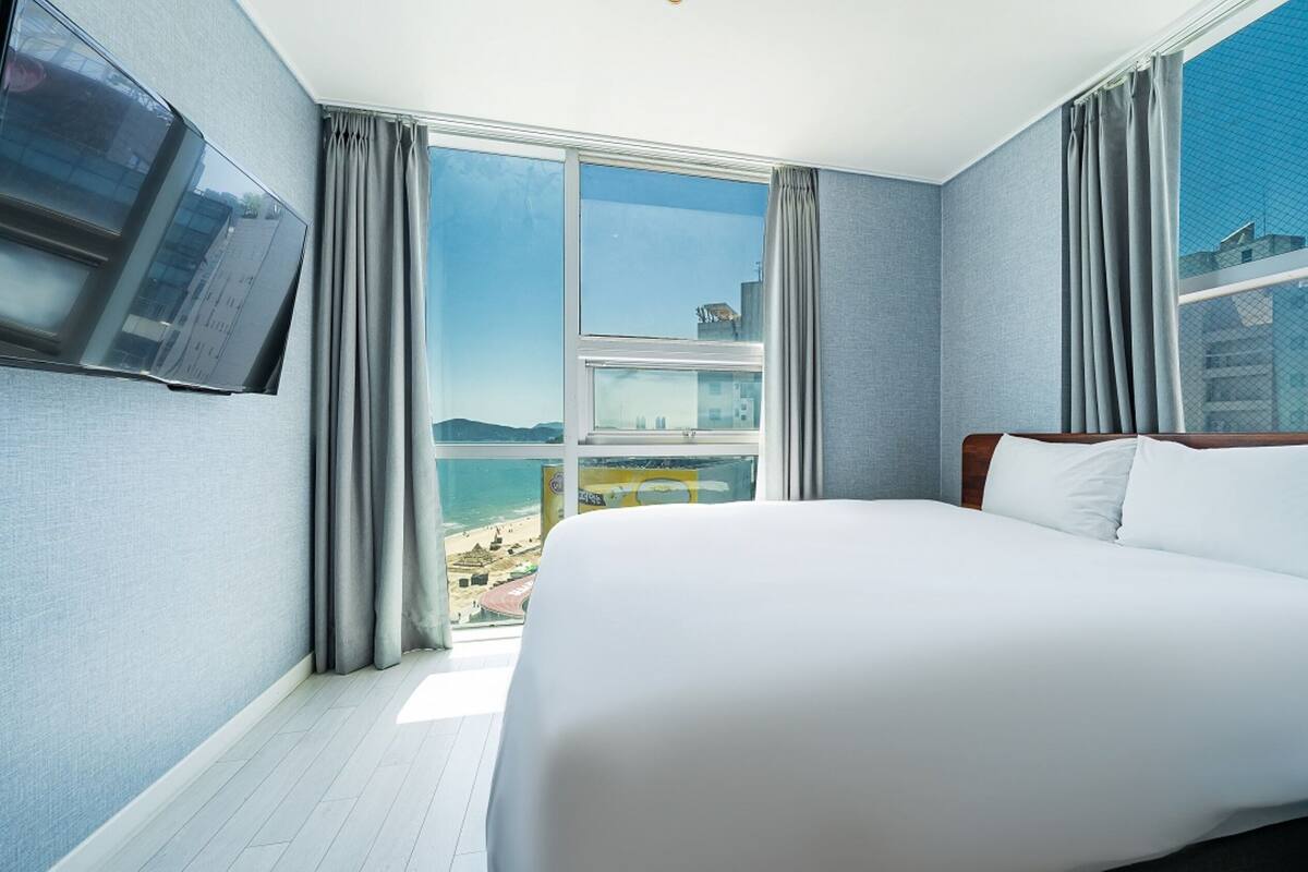 A hotel room with a large bed, a TV mounted on the wall, and a view of the beach and cityscape through the window.