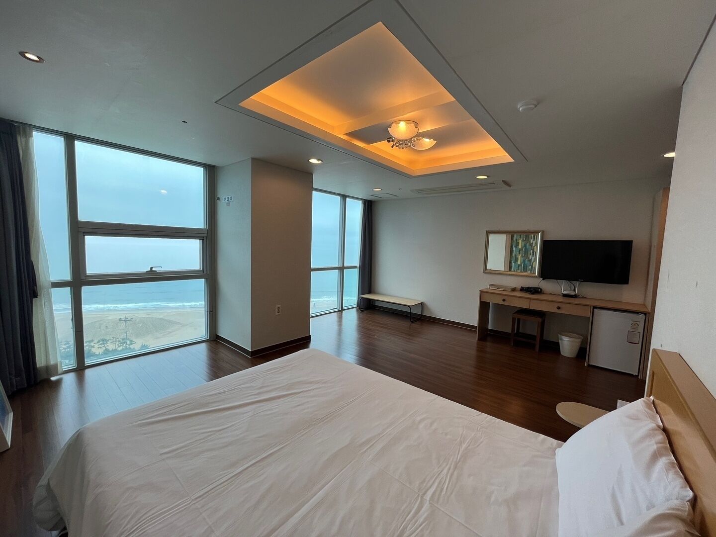 A hotel room with a large bed, a desk with a TV, and a view of the beach through large windows.