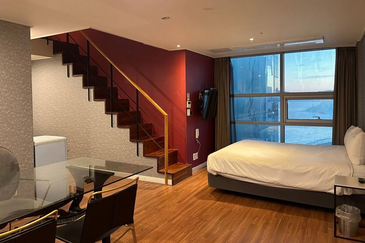 A modern hotel room with a wooden staircase, a glass dining table, a bed with white bedding, and a large window offering a city view.