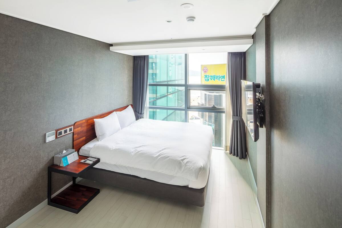 A modern hotel room with a large bed, a bedside table with a lamp, a window with curtains, and a view of the cityscape.