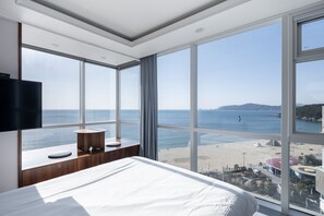 A bedroom with a large window overlooking a beach and sea, a bed, a small table, and a TV mounted on the wall. - Elysia Boutique Hotel (Busan)