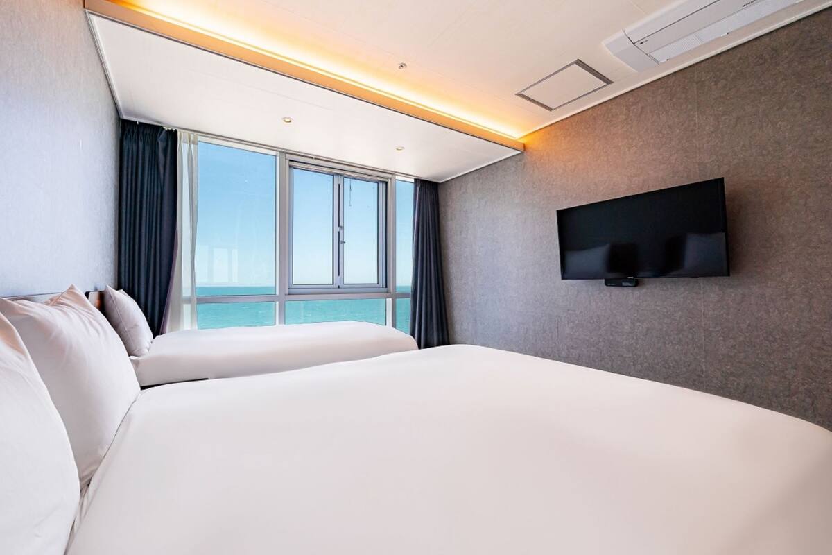 A hotel room with a large bed, a flat-screen TV mounted on the wall, and a window offering a view of the sea.