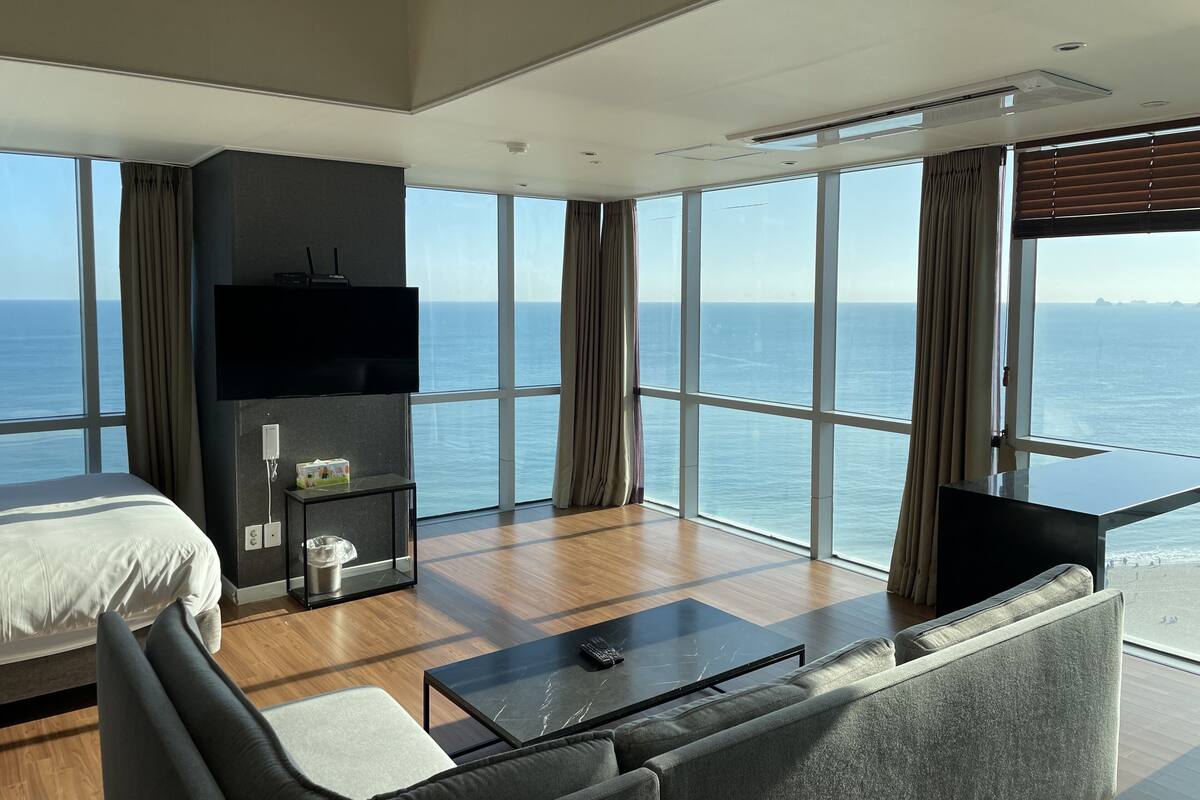 A modern hotel room with a large bed, a sofa, a small table, and a TV. The room has large windows offering a view of the sea.