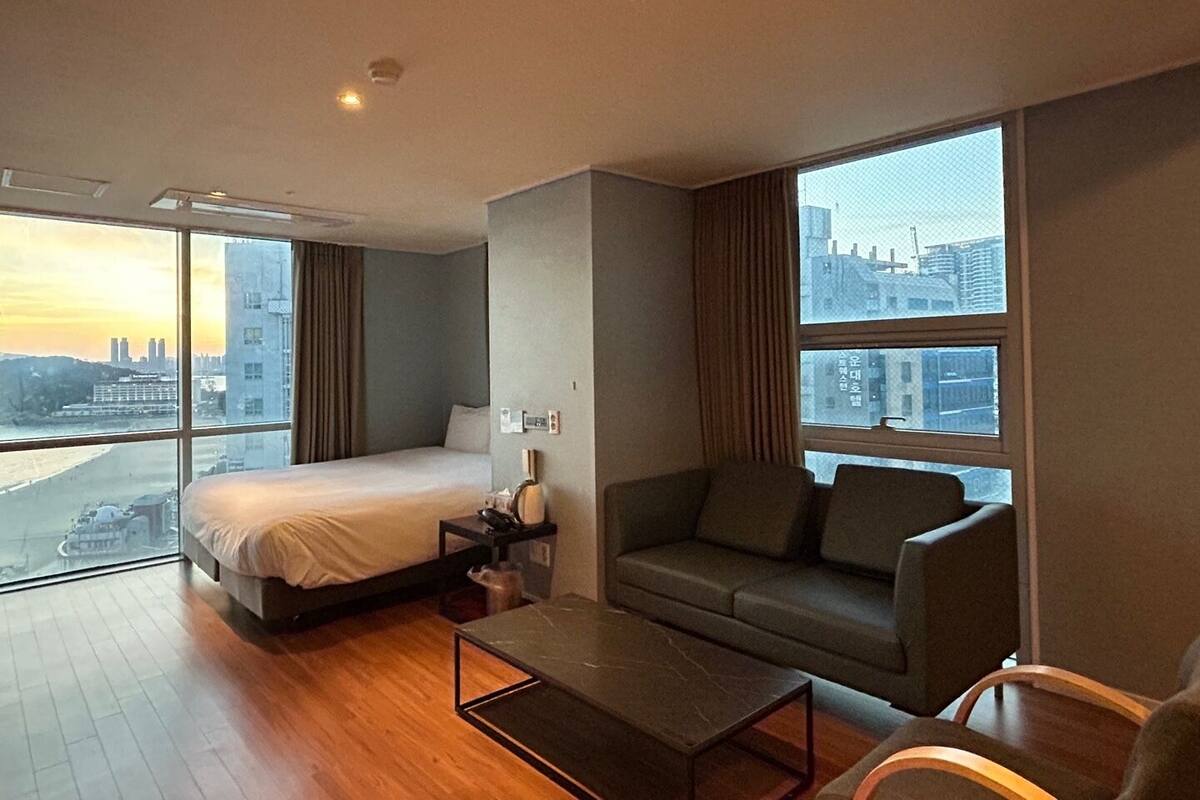 A modern hotel room with a bed, a sofa, a coffee table, and large windows offering a city view.
