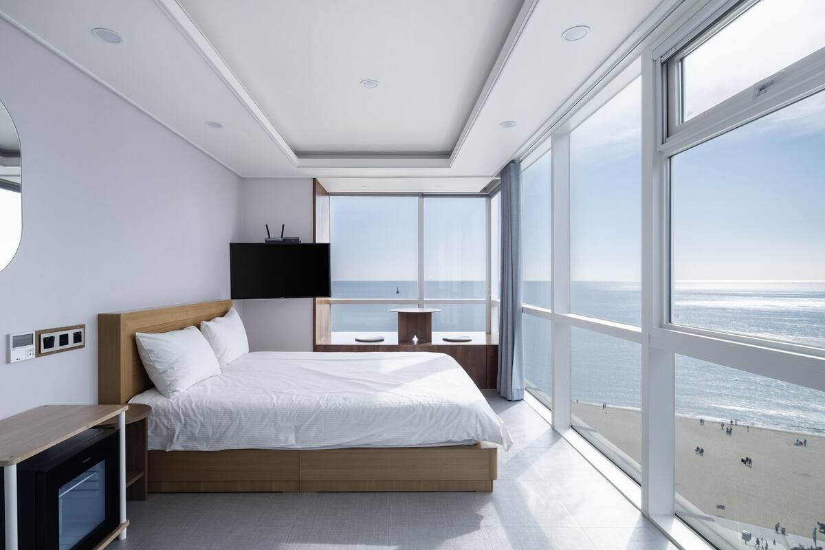 A modern bedroom with a large bed, a TV, and a view of the beach.
