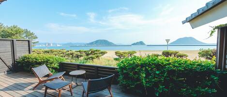 Una terraza de madera con dos sillas y una mesa, con vista a una playa y una vista despejada del mar y montañas distantes.