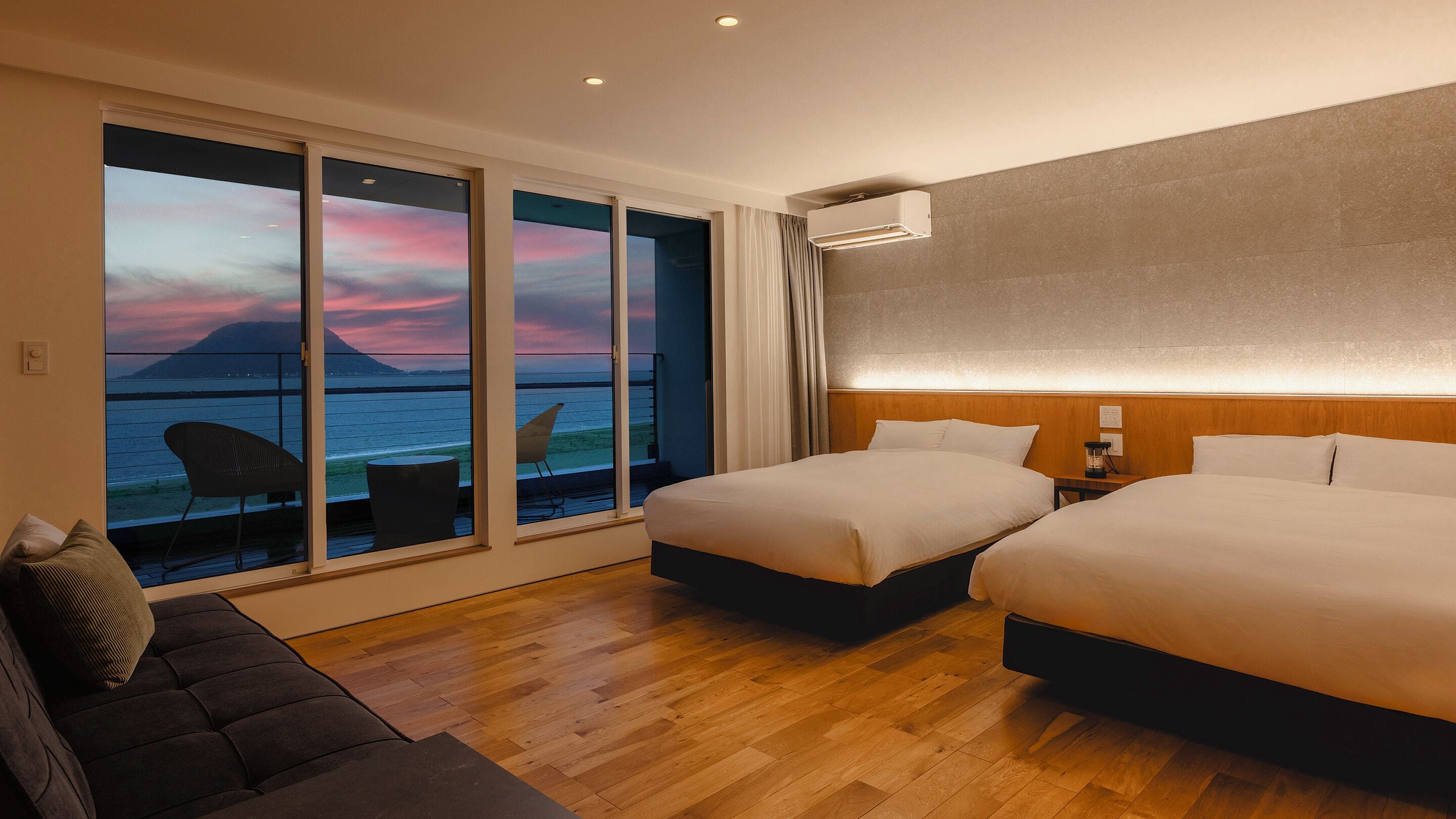 A hotel room with a large balcony, two beds, a sofa, and a view of the sea at sunset.