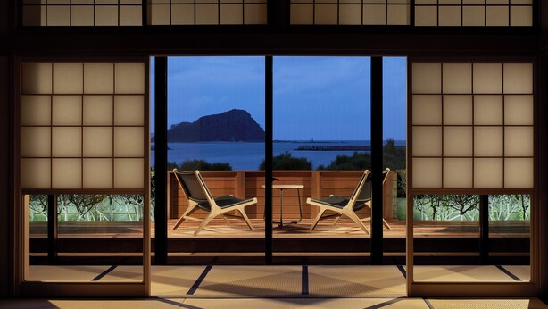 A traditional Japanese room with tatami flooring, a low table, and two chairs facing a large window overlooking a body of water and a distant mountain. - Mizuno Ryokan (Karatsu)